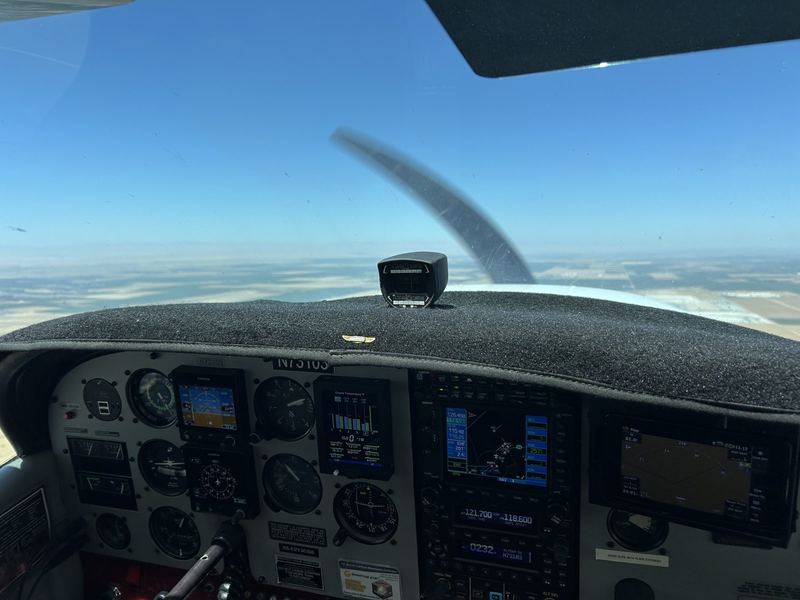 Daytime instrument panel with a Garmin G5 attitude indicator, an engine monitor, a Garmin GPS and radio stack and a moving-map display, with farmland below