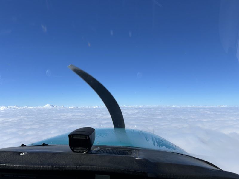 View from the cockpit of a small single-engine airplane above a solid layer of white clouds under a deep blue sky, with the propeller arc visible