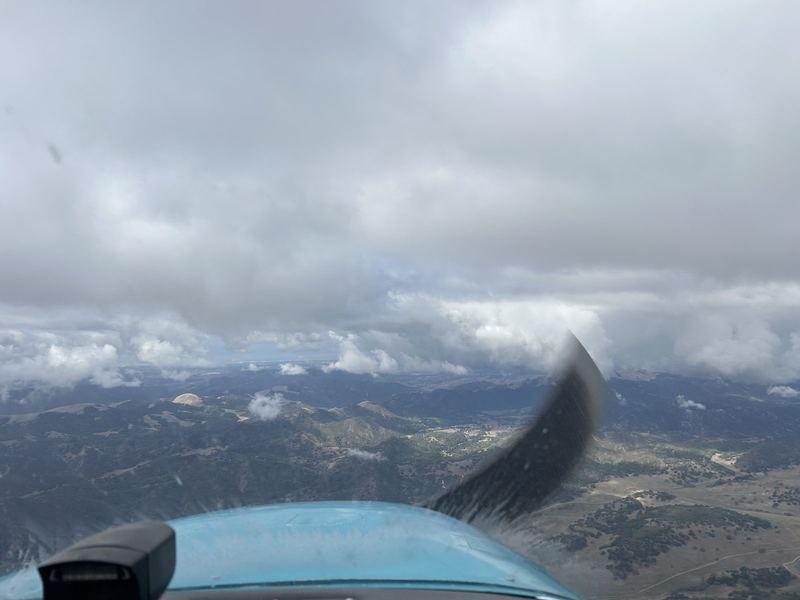Flying over tree-dotted hills beneath a broken overcast cloud layer, seen through the windshield past the propeller